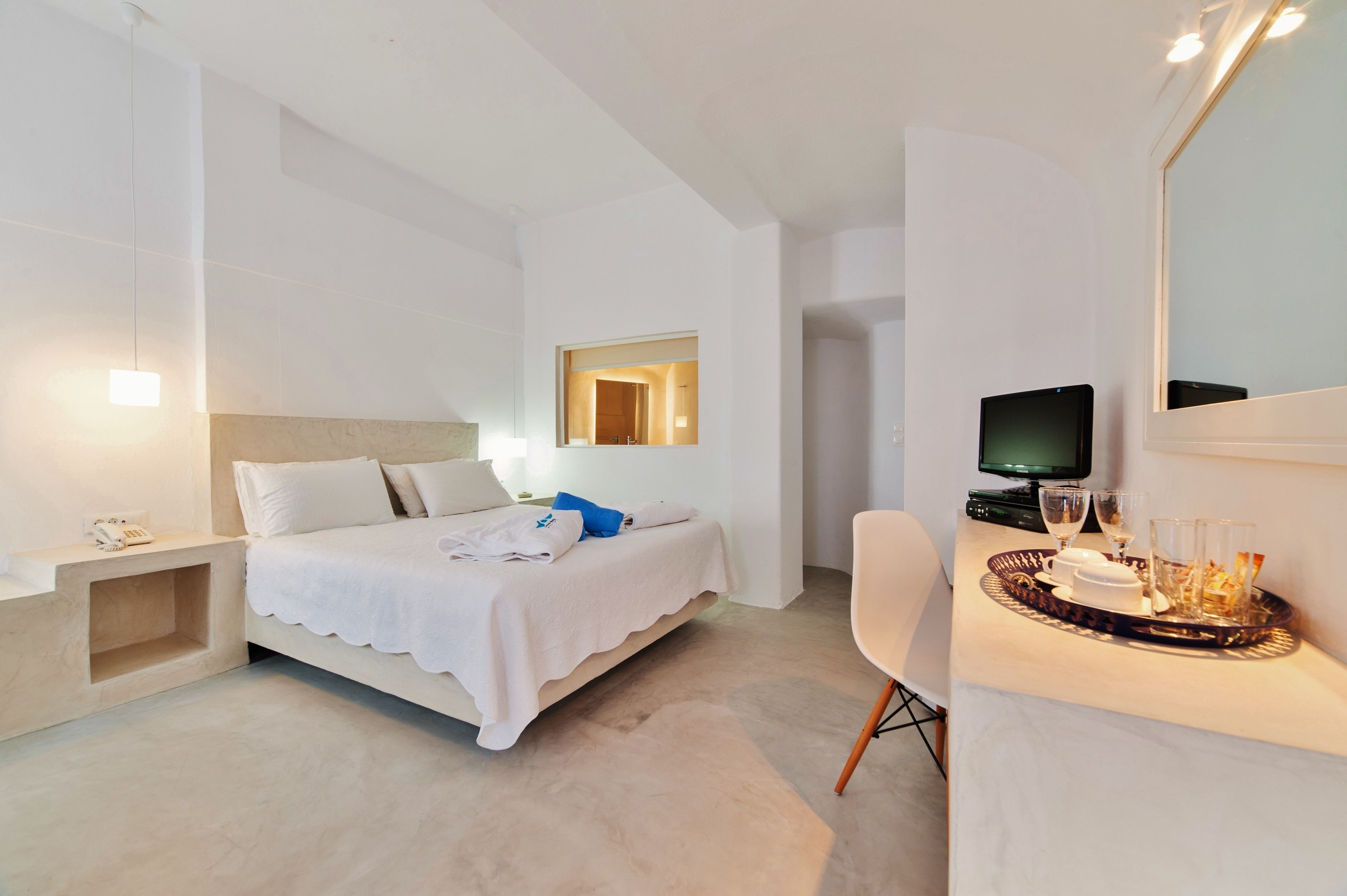 A modern hotel room with a bed, bedside table, TV, and a tray with glasses and a cup.