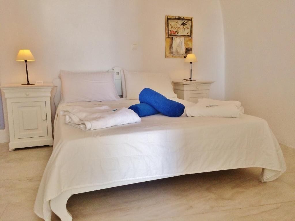 Uma cama bem arrumada com lençóis brancos e meias azuis, um criado-mudo branco com uma luminária e um relógio de parede.