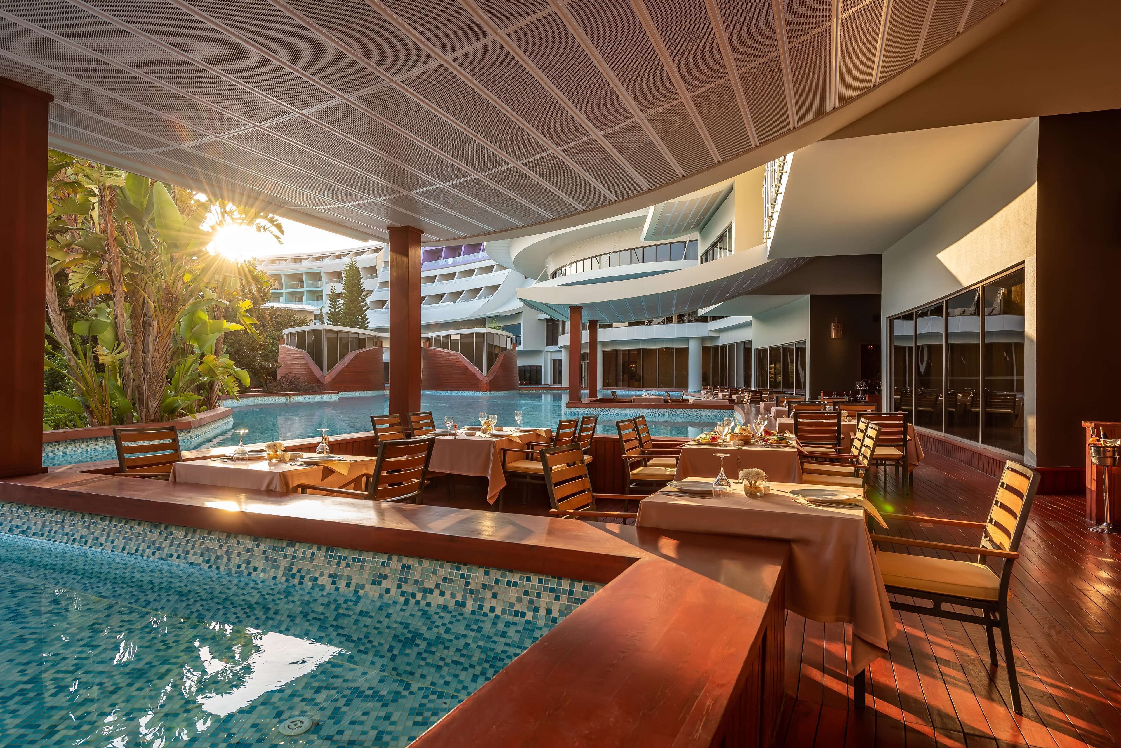 Un área de comedor junto a la piscina con mesas y sillas, una piscina y un edificio moderno al fondo.