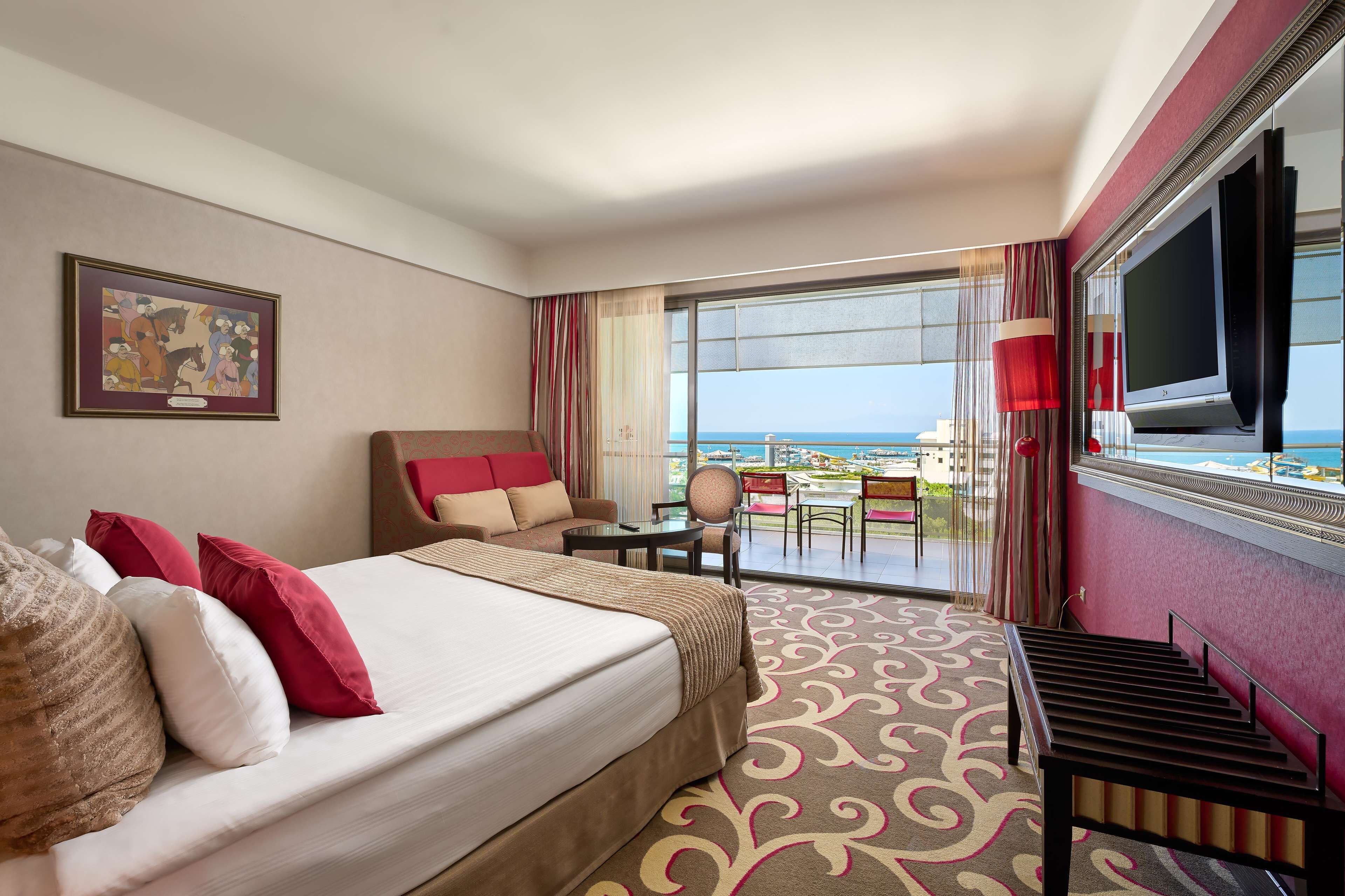 A hotel room with a large bed, a TV, a balcony with a view of the sea, and a painting on the wall.