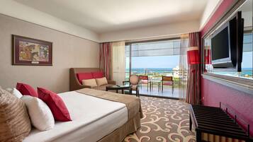 A hotel room with a large bed, a TV, a balcony with a view of the sea, and a painting on the wall.