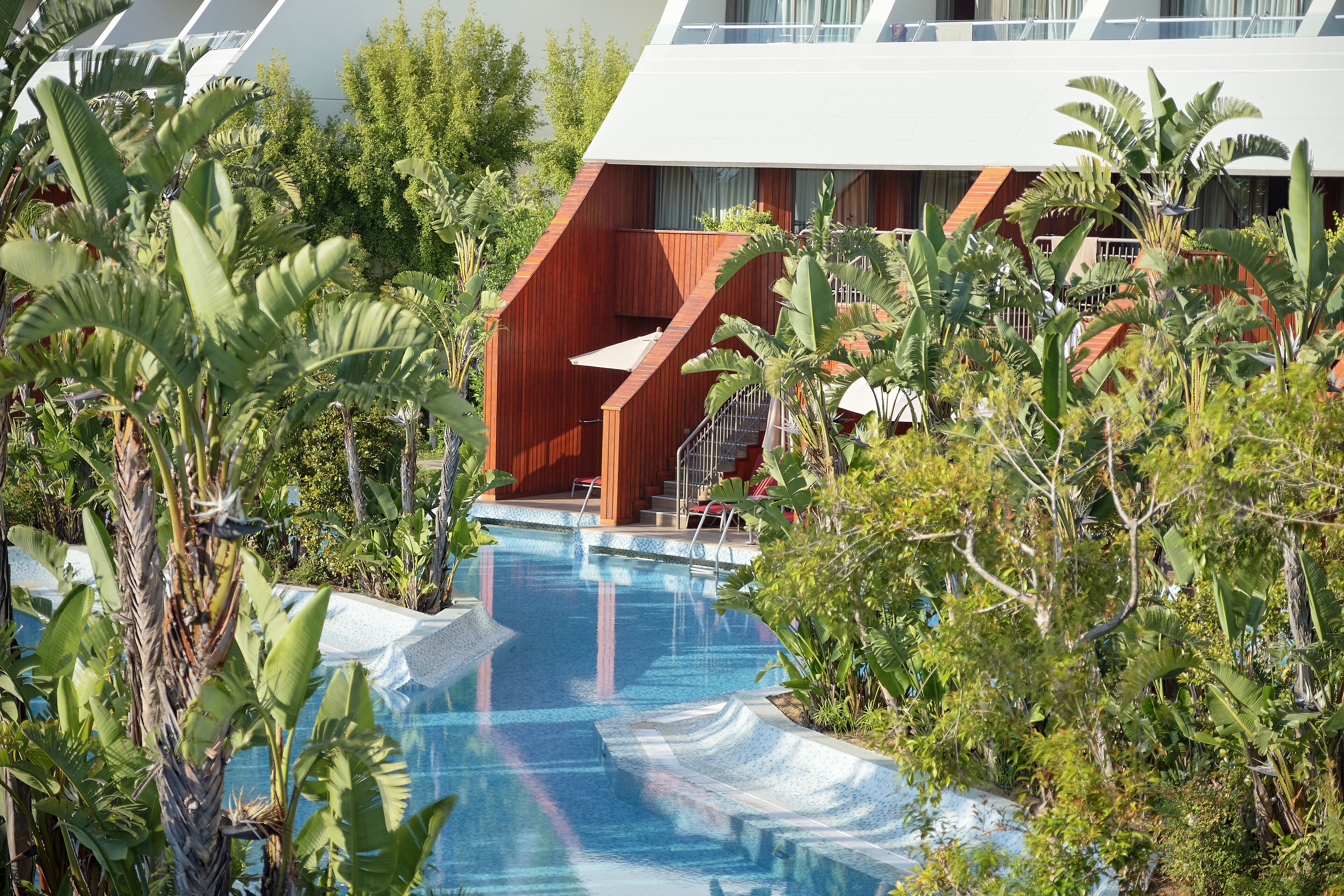 A swimming pool surrounded by lush tropical plants, with a modern building in the background.