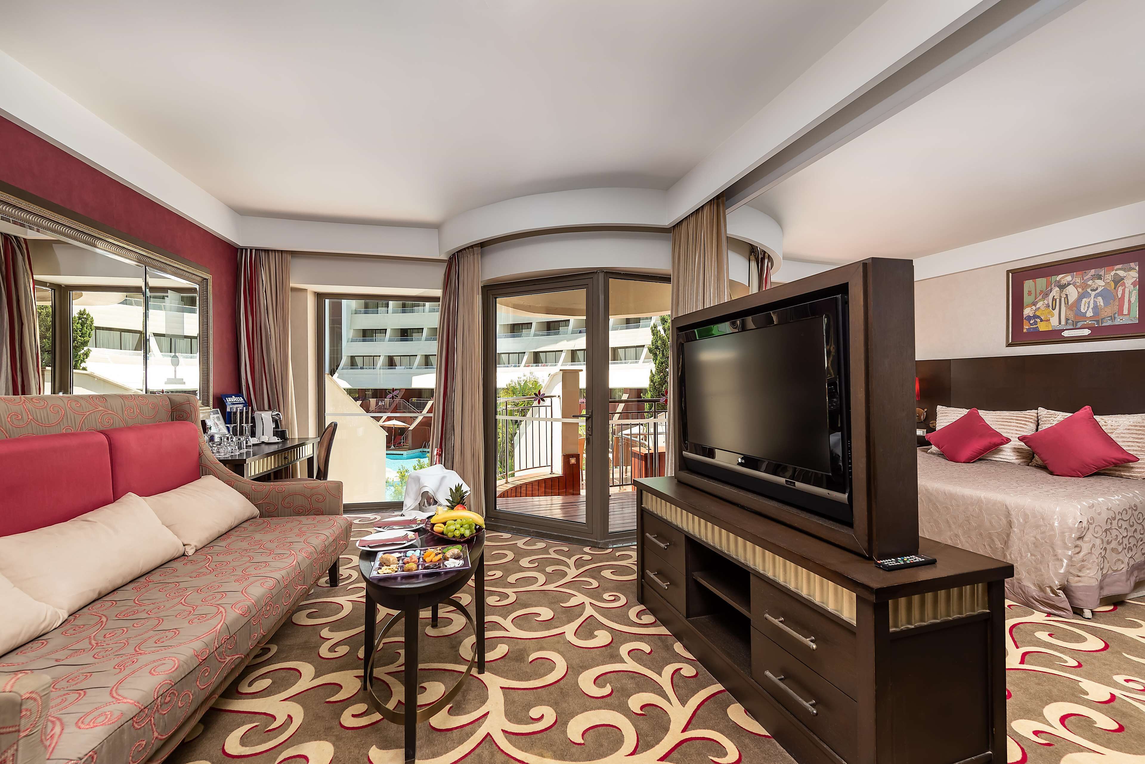 A hotel room with a sofa, a TV, a bed, a dining table with fruits, and a view of the pool and building.