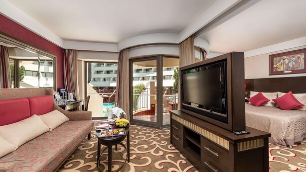 Ein Hotelzimmer mit Sofa, Fernseher, Bett, Esstisch mit Früchten sowie Blick auf den Pool und das Gebäude.