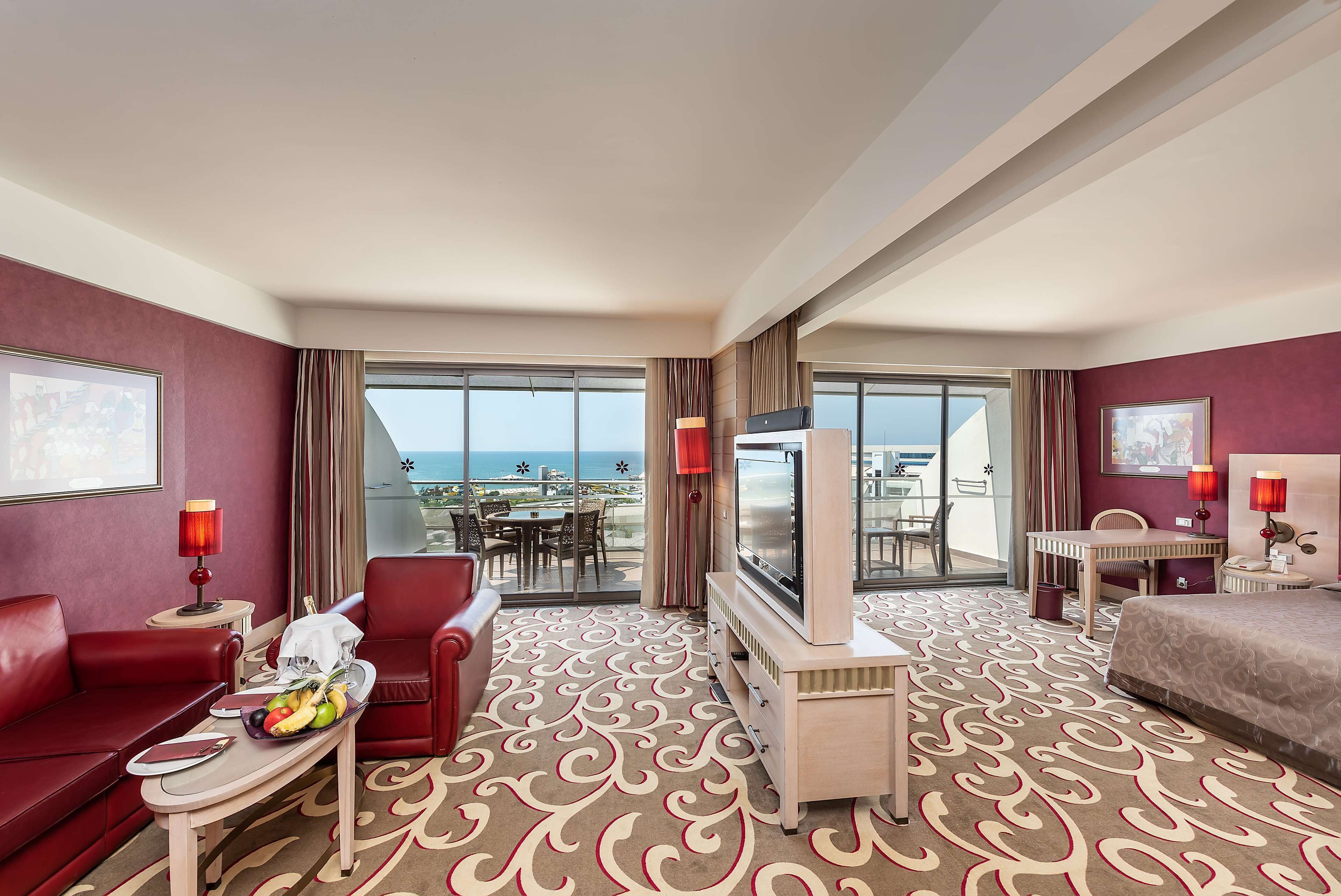 Quarto de hotel com sofá vermelho, mesa de jantar com cadeiras, televisão, cama e varanda com vista para o oceano.