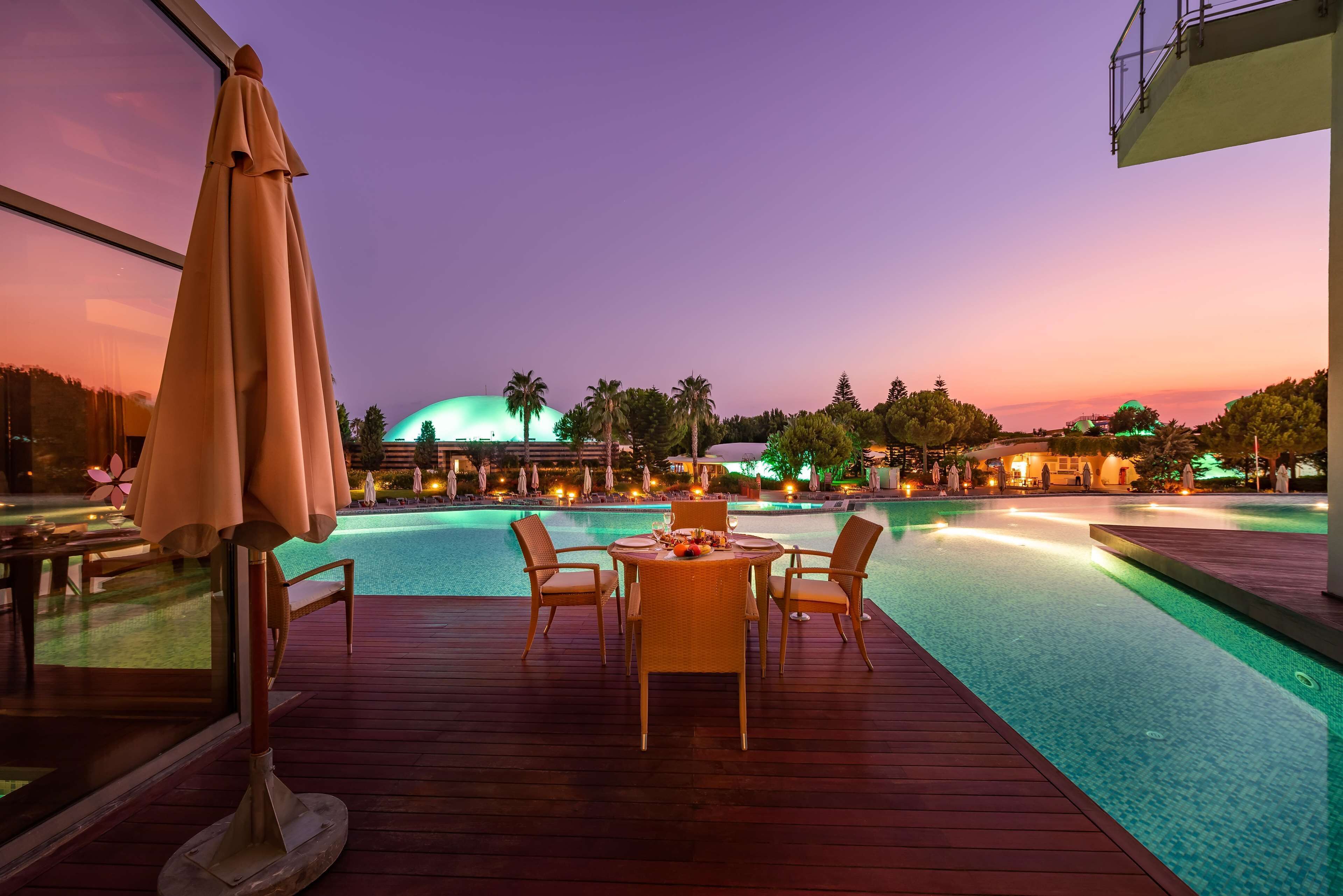 Un espace aménagé au bord de la piscine, avec une table et des chaises, un grand parasol et une vue sur un bâtiment doté d’un dôme et bordé de palmiers.