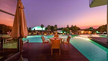 Un espace aménagé au bord de la piscine, avec une table et des chaises, un grand parasol et une vue sur un bâtiment doté d’un dôme et bordé de palmiers.