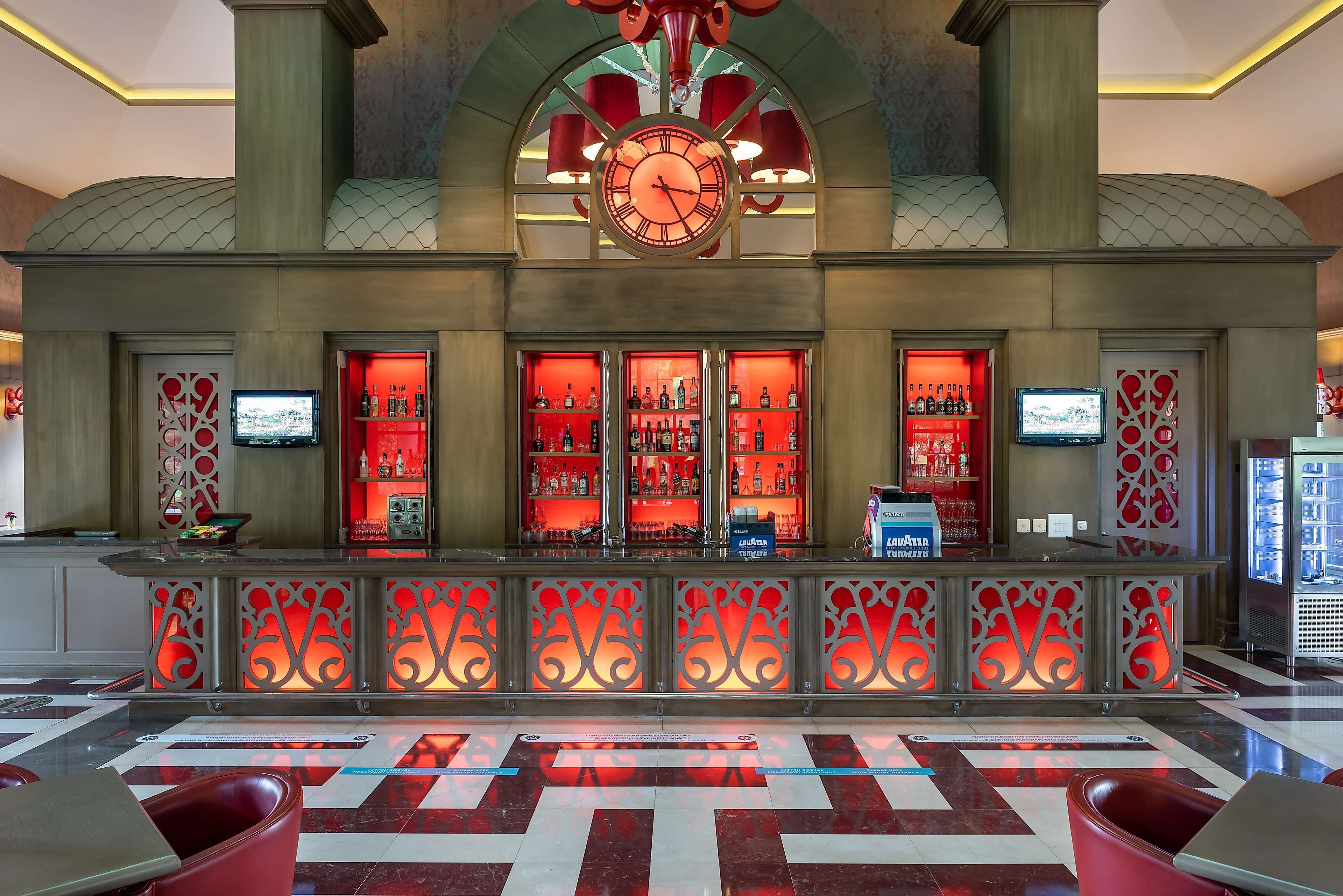 Un vestíbulo con un reloj grande, una zona de bar con iluminación roja y un mostrador de recepción.