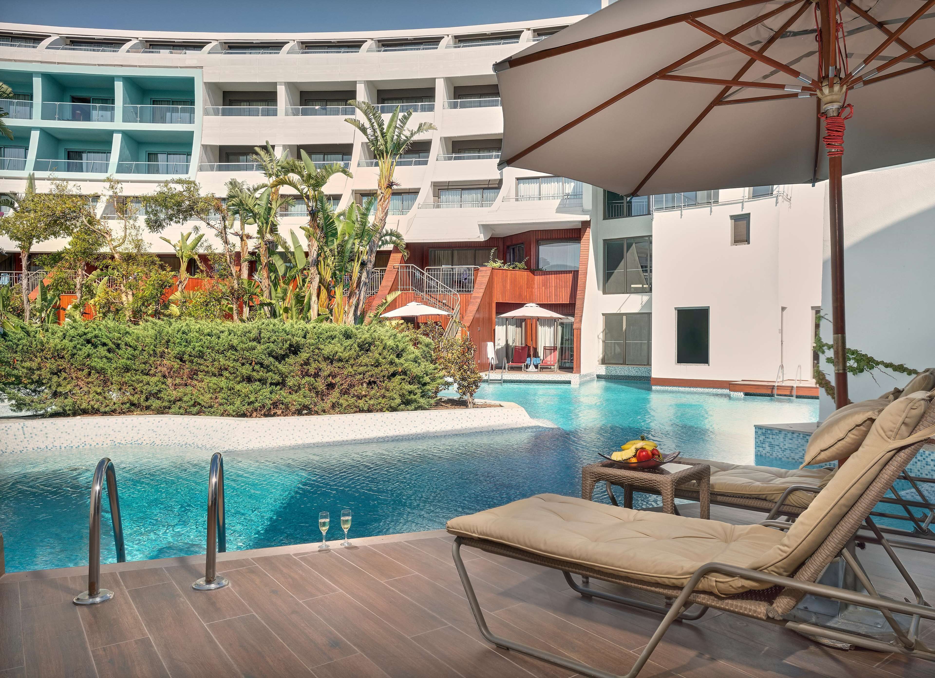 A poolside area with lounge chairs and a pool, surrounded by greenery and a multi-story building.