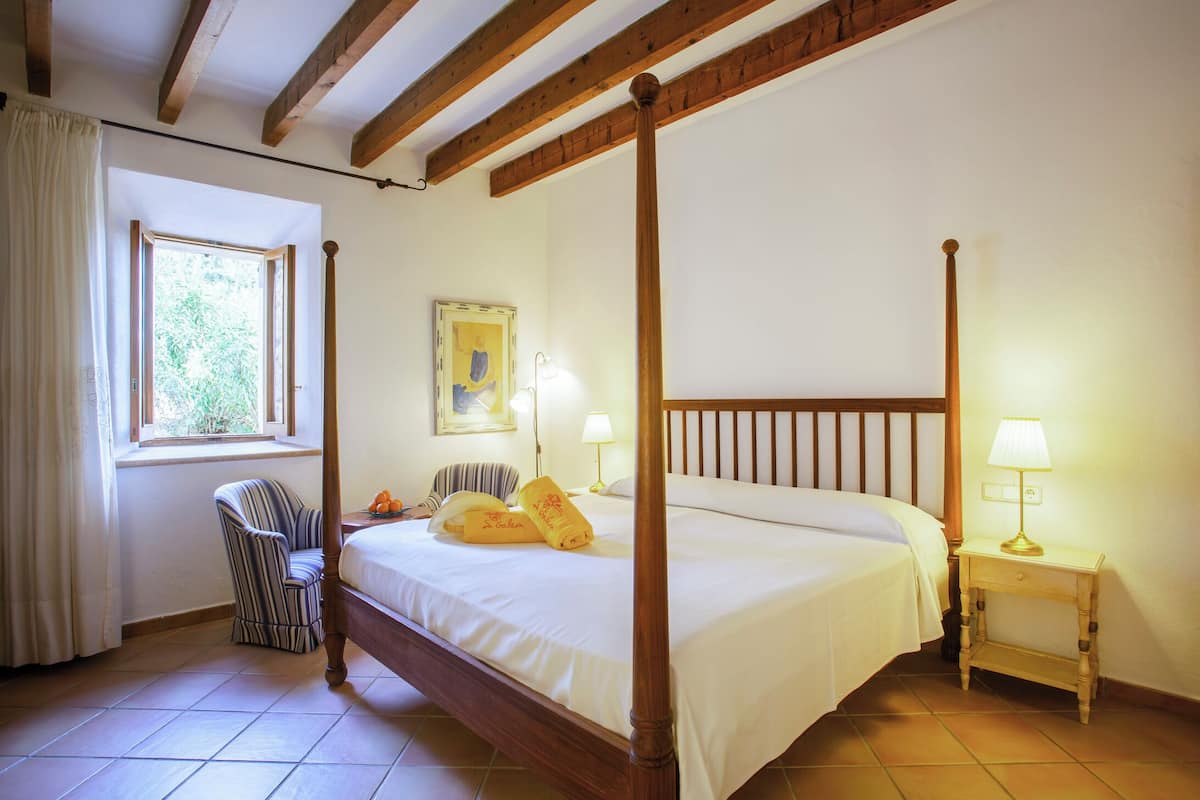 Una cama con dosel y cabecera de madera, una mesita de noche con lámpara, una silla y una ventana con cortinas.