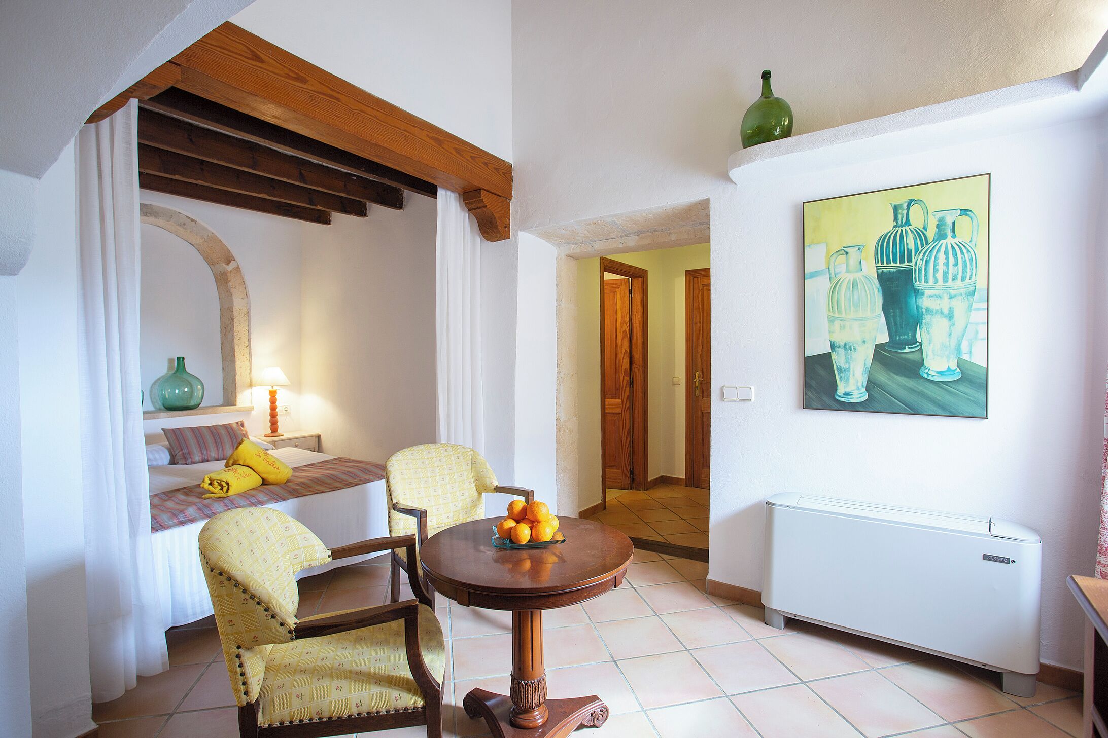 Una habitación con una cama, dos sillas, una mesa con fruta, un radiador y un cuadro en la pared.