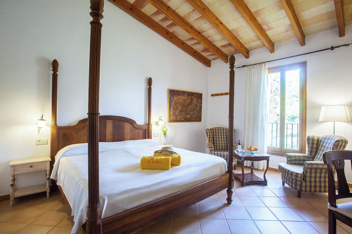Una cama con dosel con un cabecero de madera, una mesita de noche, una silla, una lámpara y una ventana con cortinas.