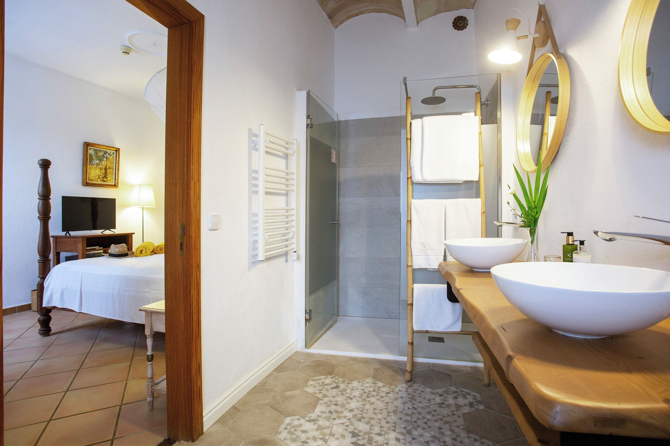 A bathroom with a shower, two sinks, a mirror, and a bed in the adjacent room.