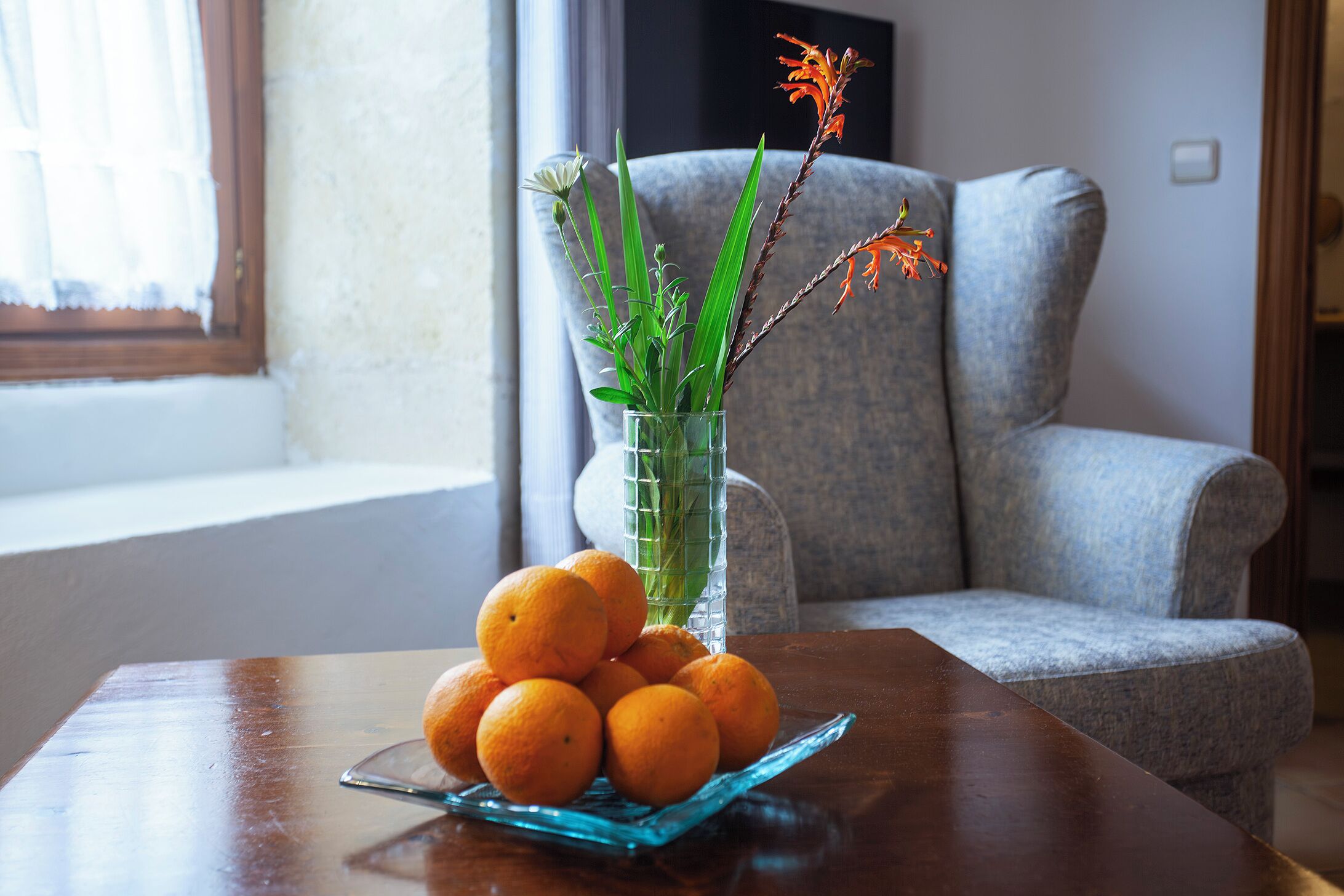 A table with a glass vase containing plants and a bowl of oranges.