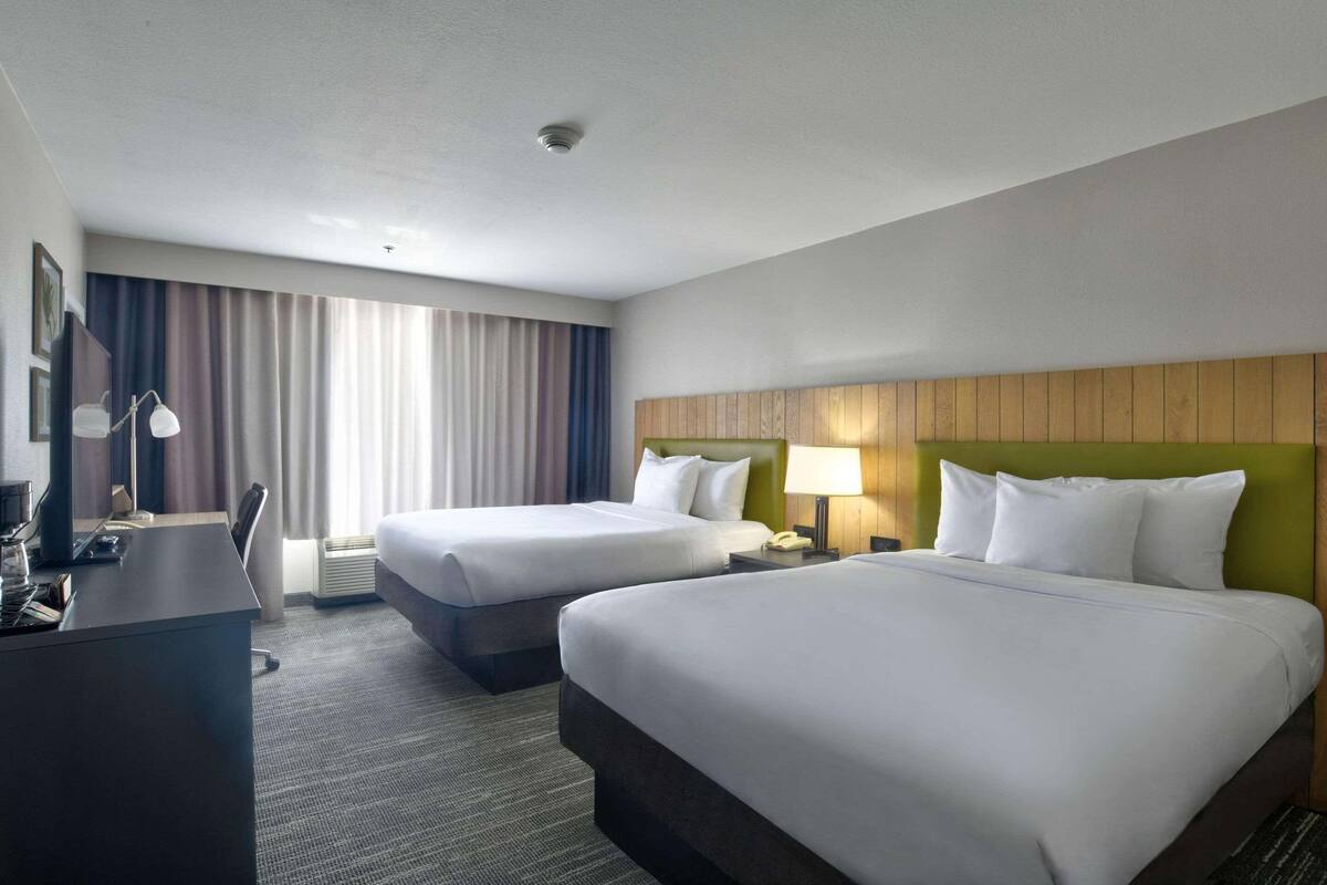 Habitación de hotel con dos camas, un escritorio, una silla, una lámpara y una ventana con cortinas.