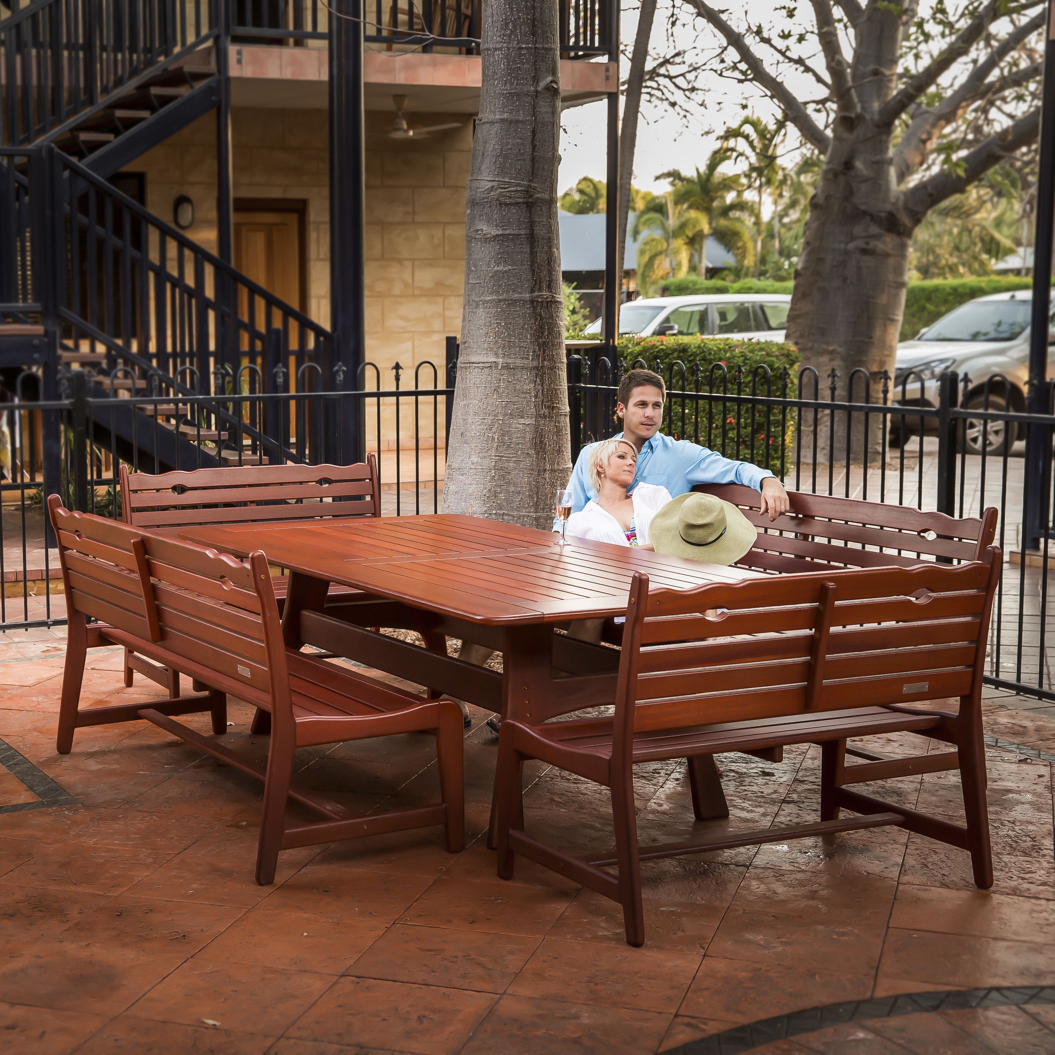 Une table et des bancs en bois, installés à l’extérieur, avec un sol carrelé, une clôture en métal et un bâtiment avec un escalier en arrière-plan.
