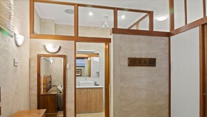 Une chambre à coucher avec un lit, une table de chevet, une commode en bois et une salle de bain visible à travers une porte ouverte.