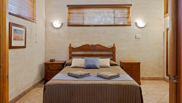 Un dormitorio ordenado con una cama de madera, dos mesitas de noche, un buró, una ventana con persianas y una lámpara fijada a la pared.