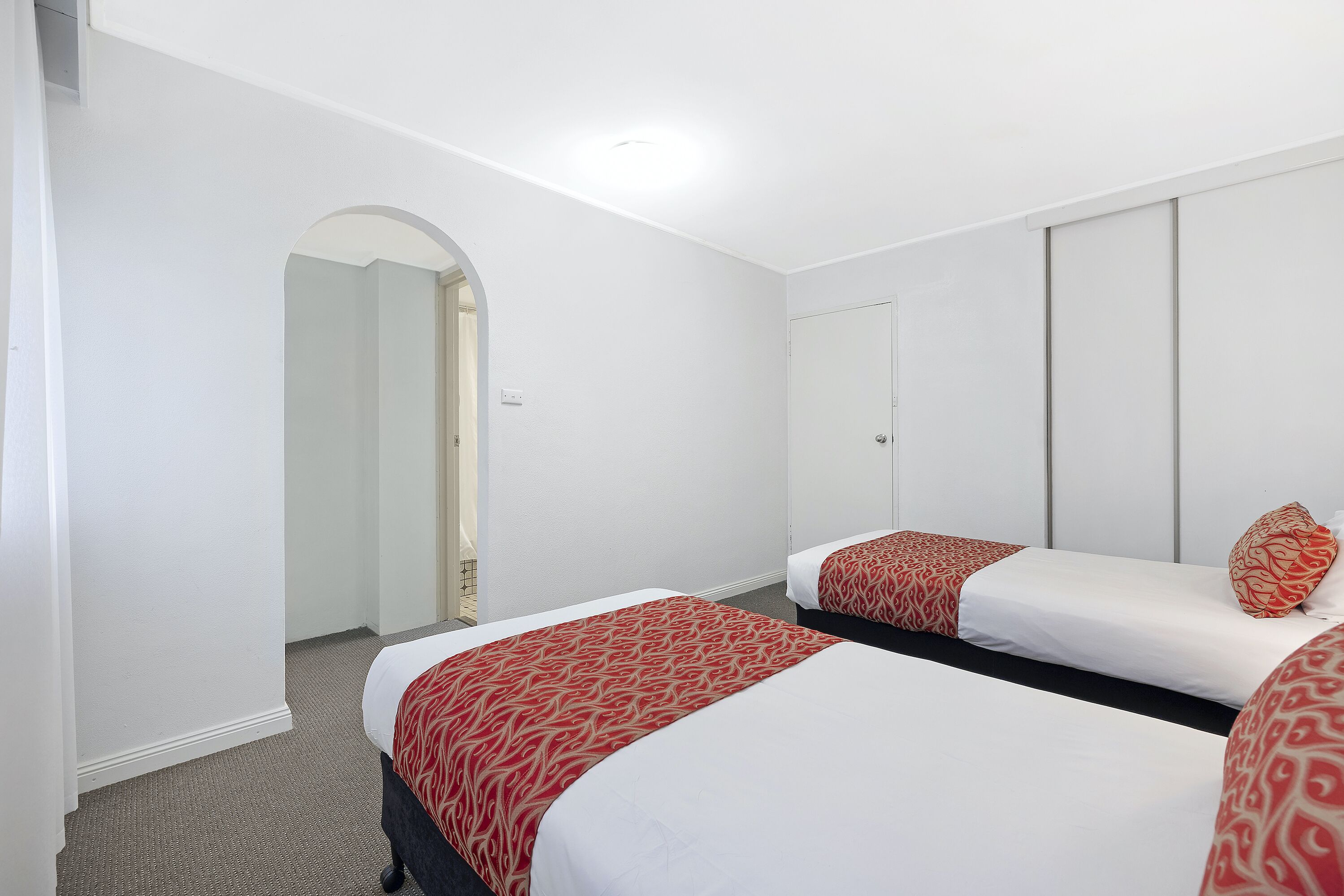 A hotel room with two single beds, white walls, a door, and a carpeted floor.