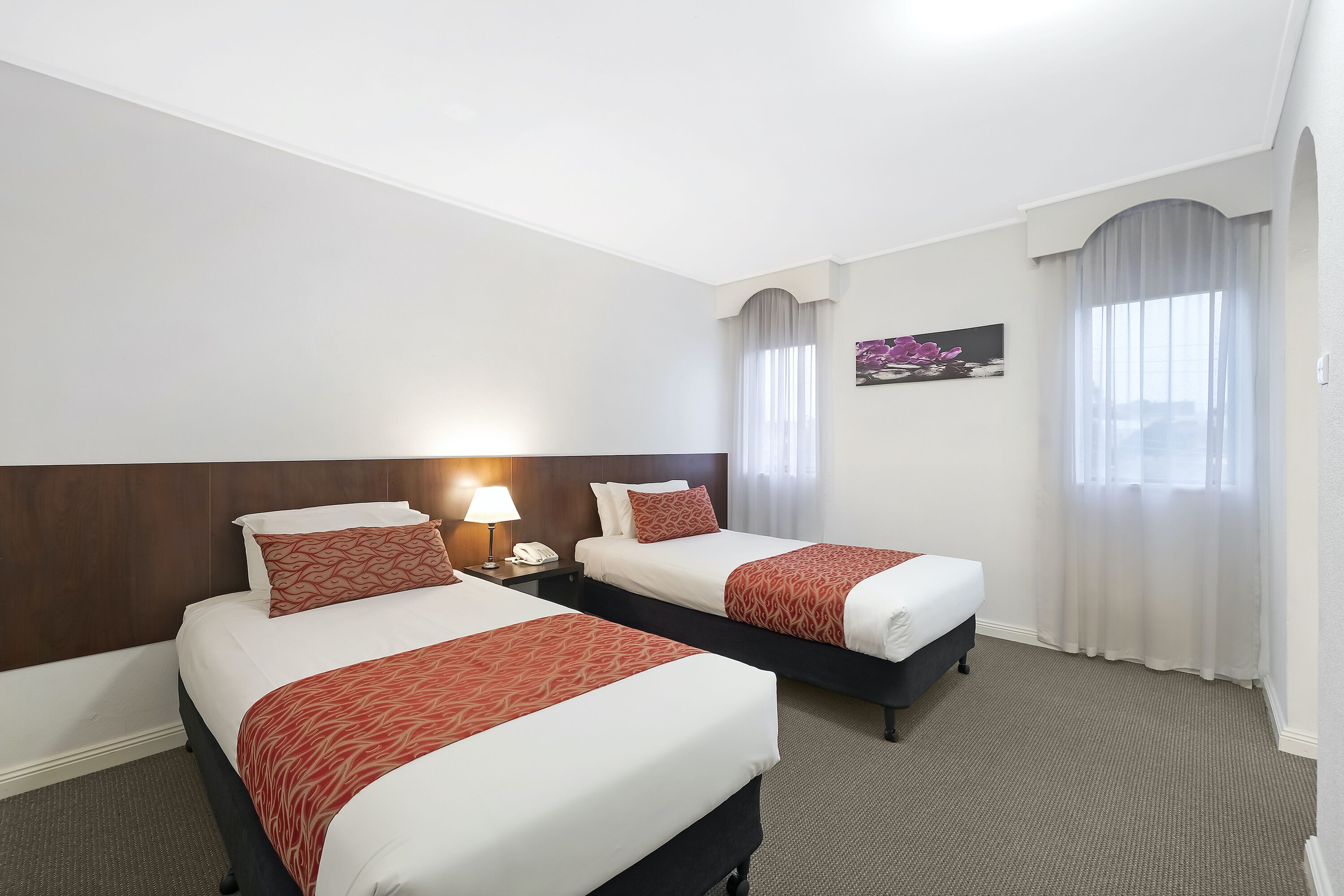 Une chambre d’hôtel avec deux lits, une table de chevet avec une lampe, un tableau au mur et une fenêtre avec des rideaux.
