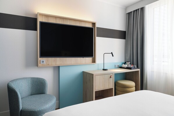A hotel room with a bed, a desk with a lamp, a television, a chair, and a small stool. - Spark by Hilton Essen City Centre (Essen)