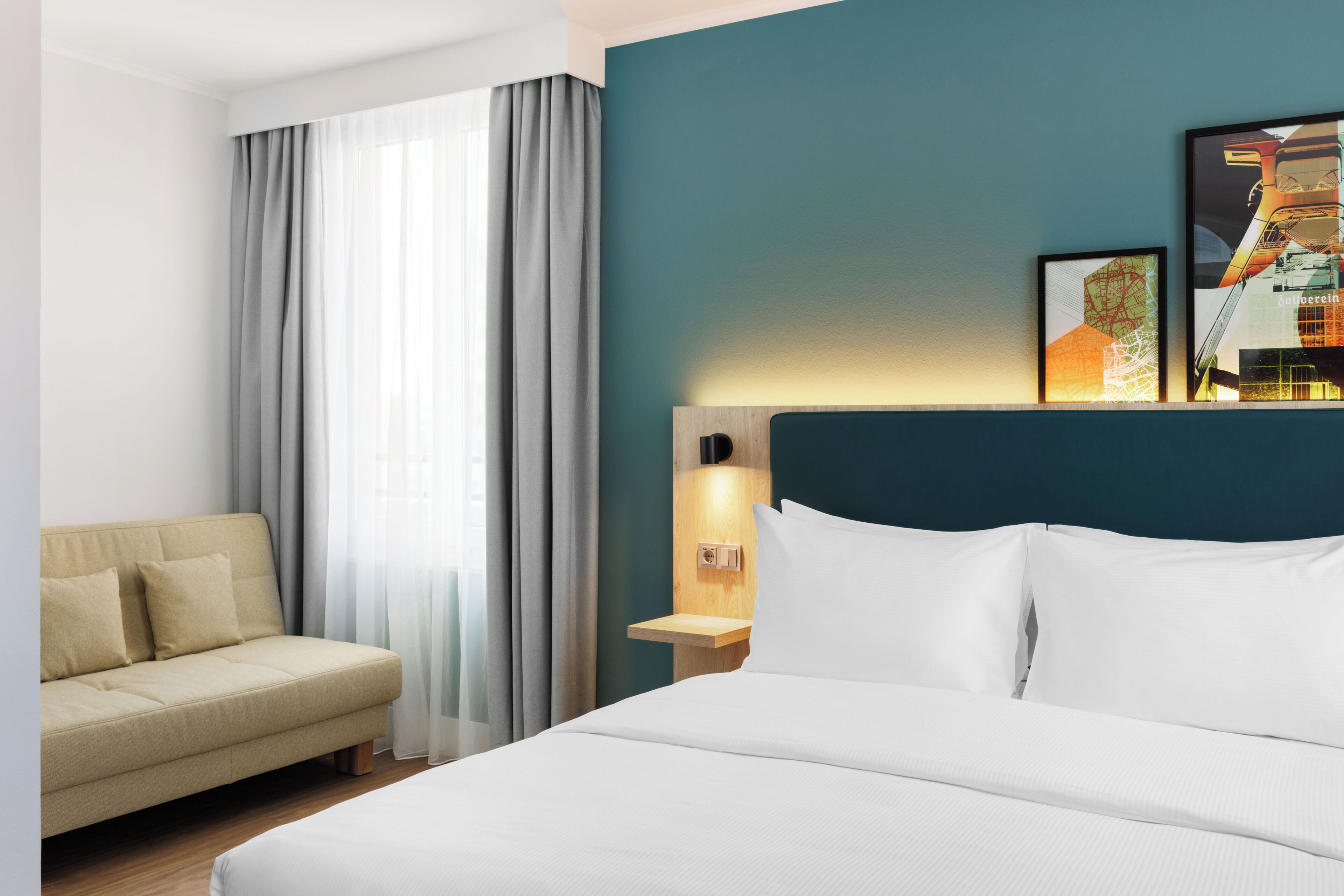 A hotel room with a bed, a sofa, a nightstand with a lamp, and a wall with framed pictures.