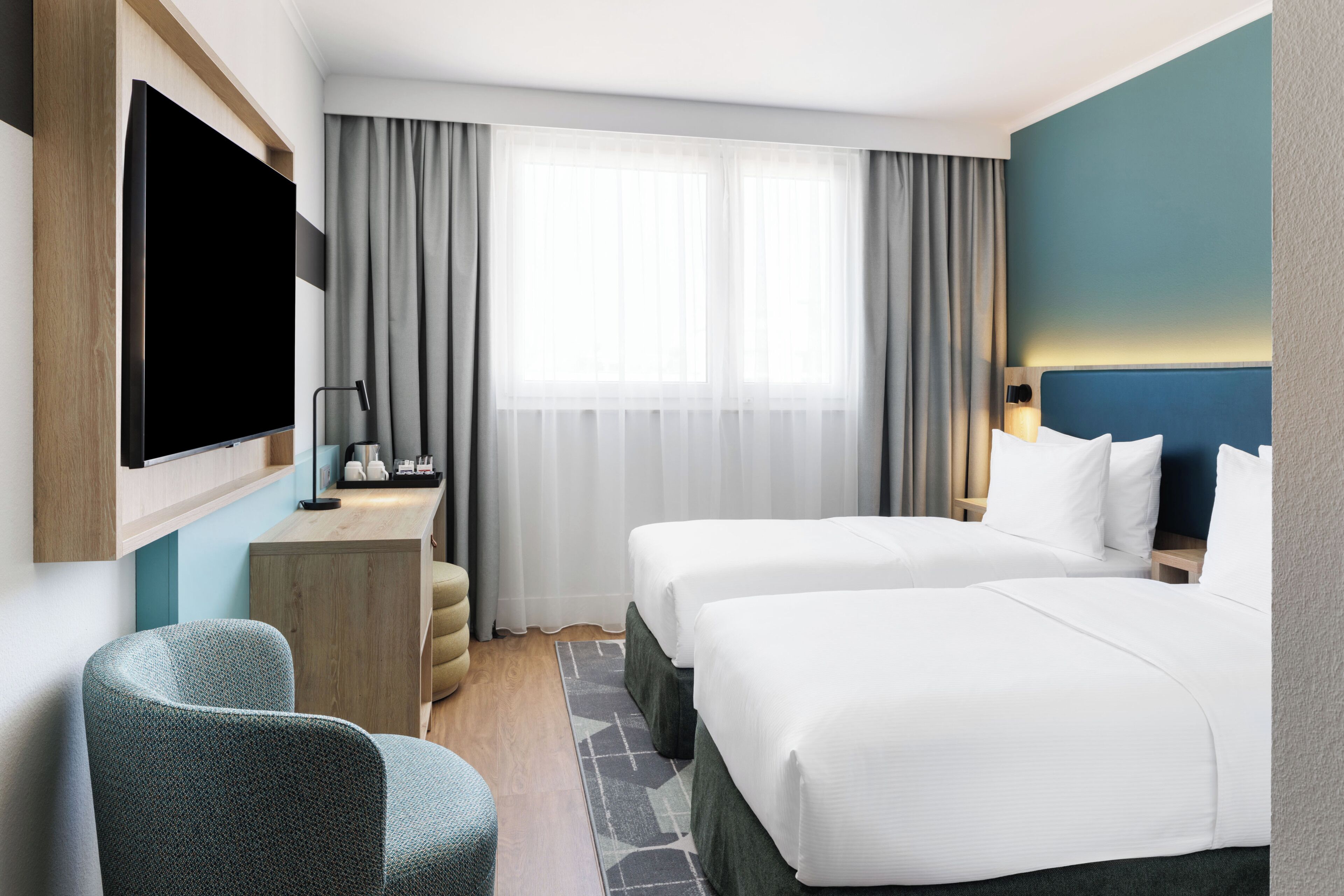 Habitación de hotel con dos camas, un televisor montado en la pared, un escritorio con lámpara y una ventana con cortinas.