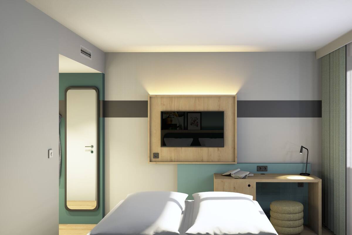 A modern hotel room with a bed, a bedside table with a lamp, a mirror, and a small wooden cabinet with a TV.