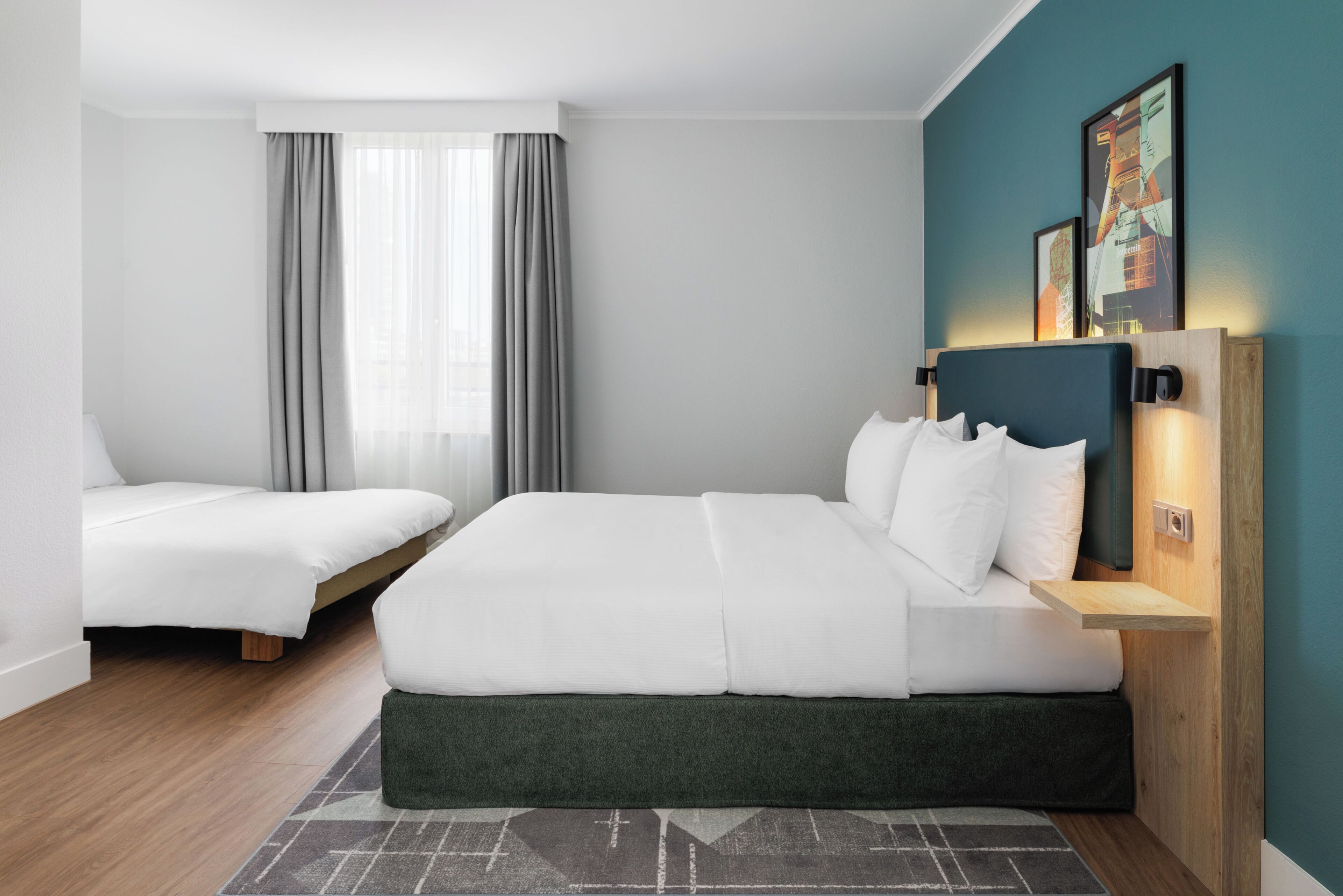Ein Hotelzimmer mit zwei Betten, einem hölzernen Kopfteil, einem Nachttisch, einer Wandlampe und einem Fenster mit Vorhängen.
