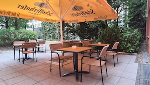 Een terras met tafels en stoelen, overdekt met gele parasols, omringd door groen.