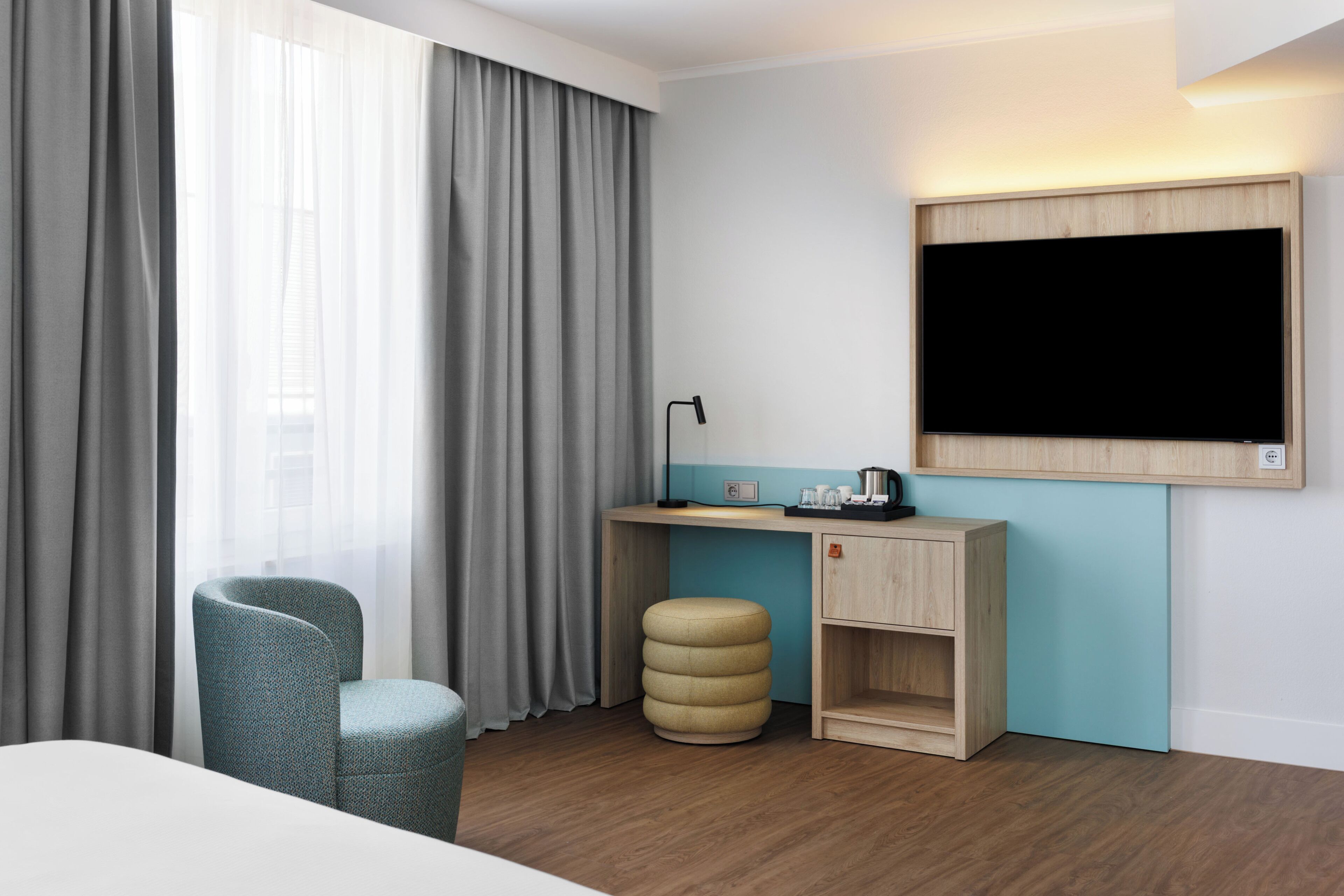 Ein Hotelzimmer mit einem Bett, einem Schreibtisch mit Lampe, einem Stuhl, einem kleinen Hocker und einem an der Wand befestigten Fernseher.