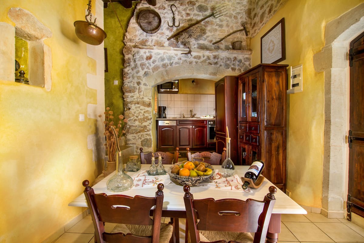 A dining area with a stone archway leading to a kitchen, wooden chairs, a table with a fruit bowl, and a bottle of wine.