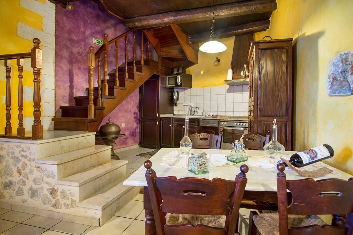 A dining area with a table set for four, wooden chairs, a staircase, and a kitchen in the background.