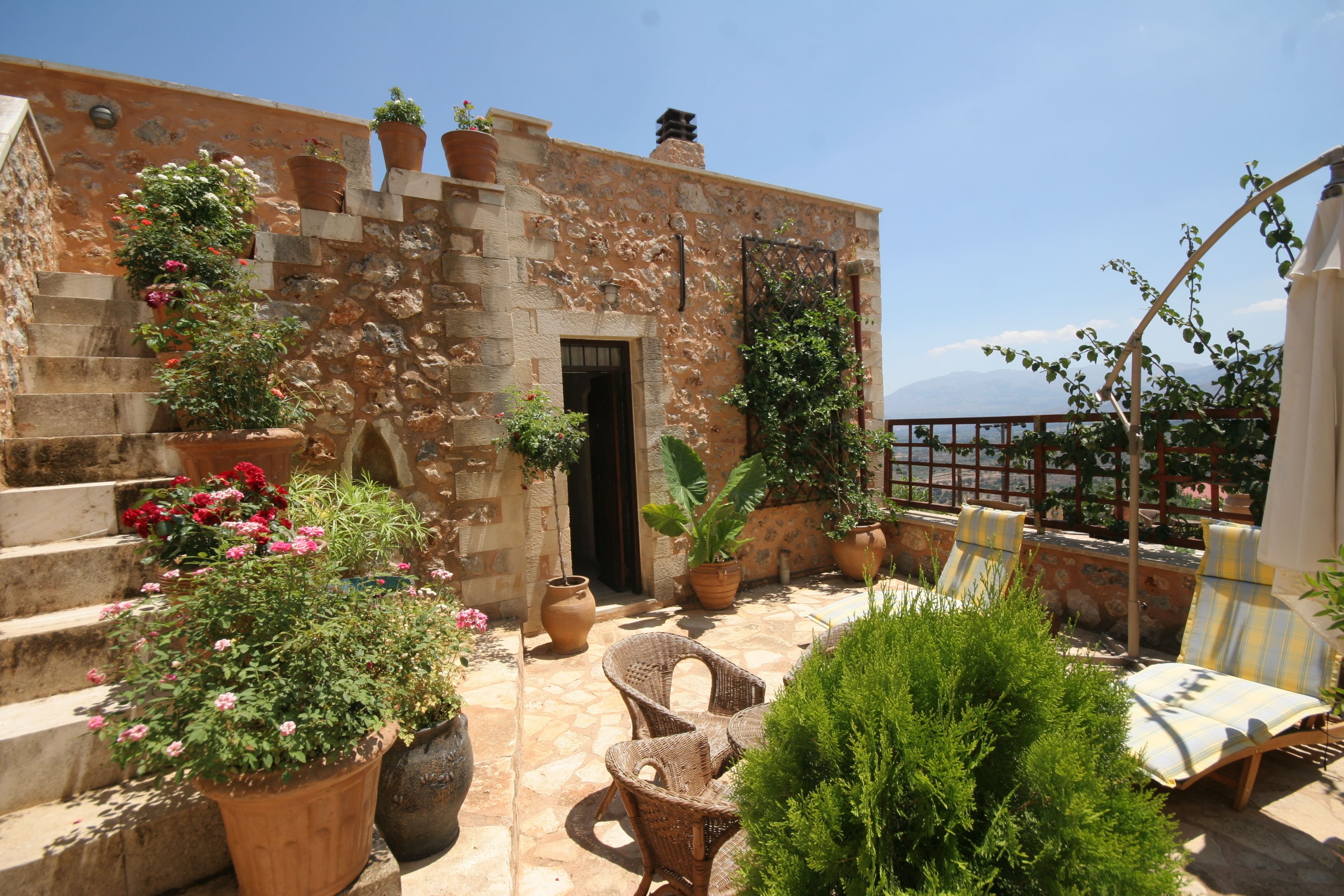 Un bâtiment en pierre avec un escalier menant à une porte, entouré de plantes en pot et de meubles en osier.