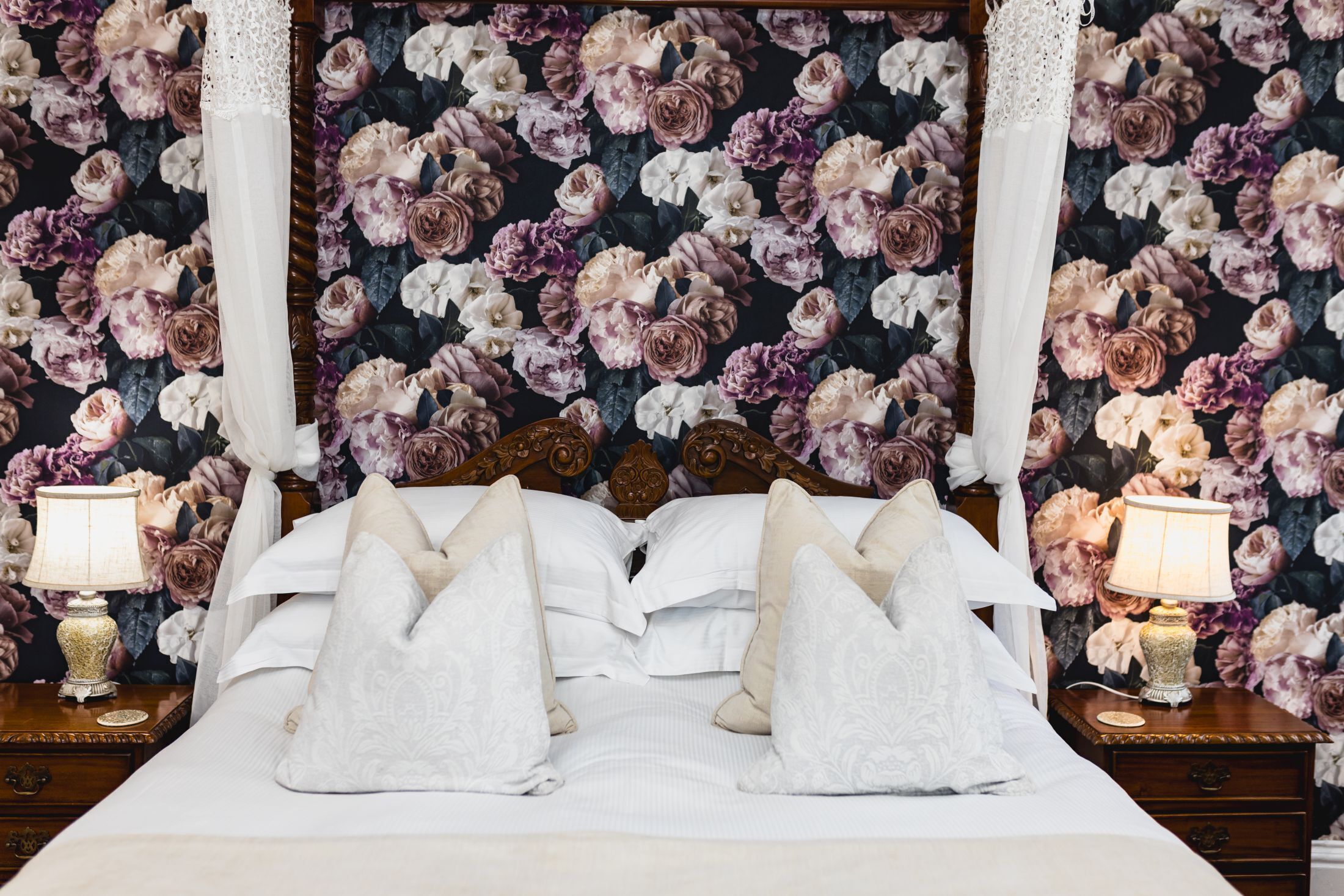 Una cama con ropa blanca y múltiples almohadas, flanqueada por dos mesitas de noche elaboradas con lámparas, sobre un fondo de papel tapiz floral.