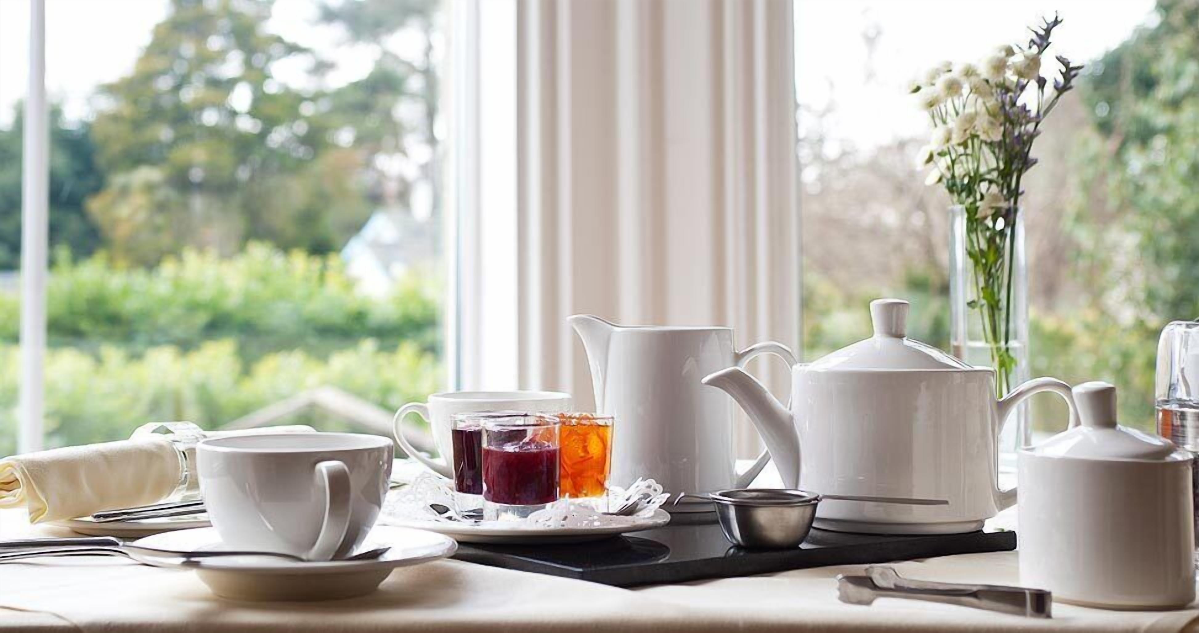A table set for tea with white teacups, a teapot, and a pitcher, with a window view of greenery.