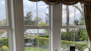 A dining area with a view of a garden through large windows, a table set with white tableware, and a menu stand. - Wheatlands Lodge - Bed & Breakfast (Windermere)