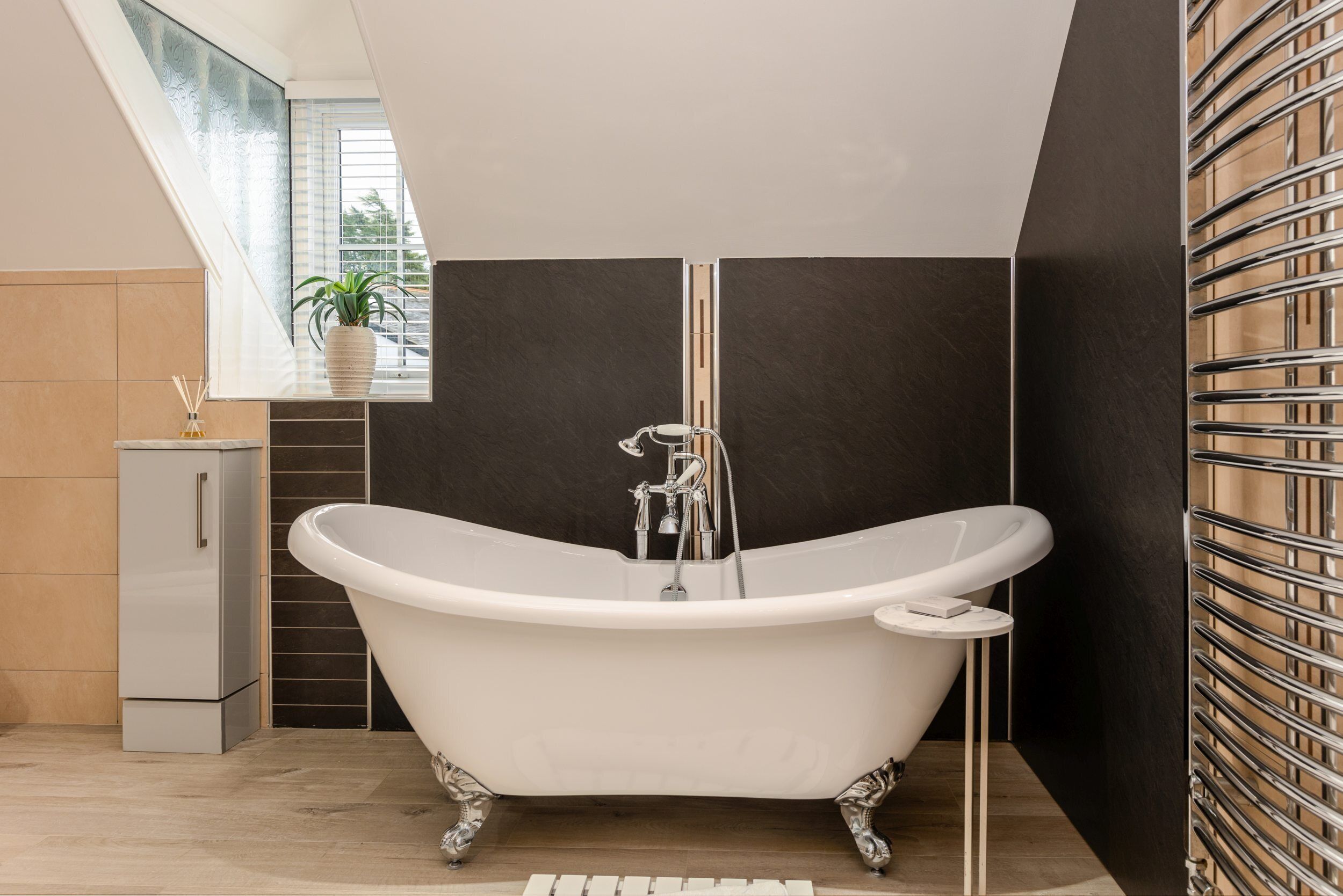 A modern bathroom with a freestanding bathtub, a small cabinet, and a window with a plant.