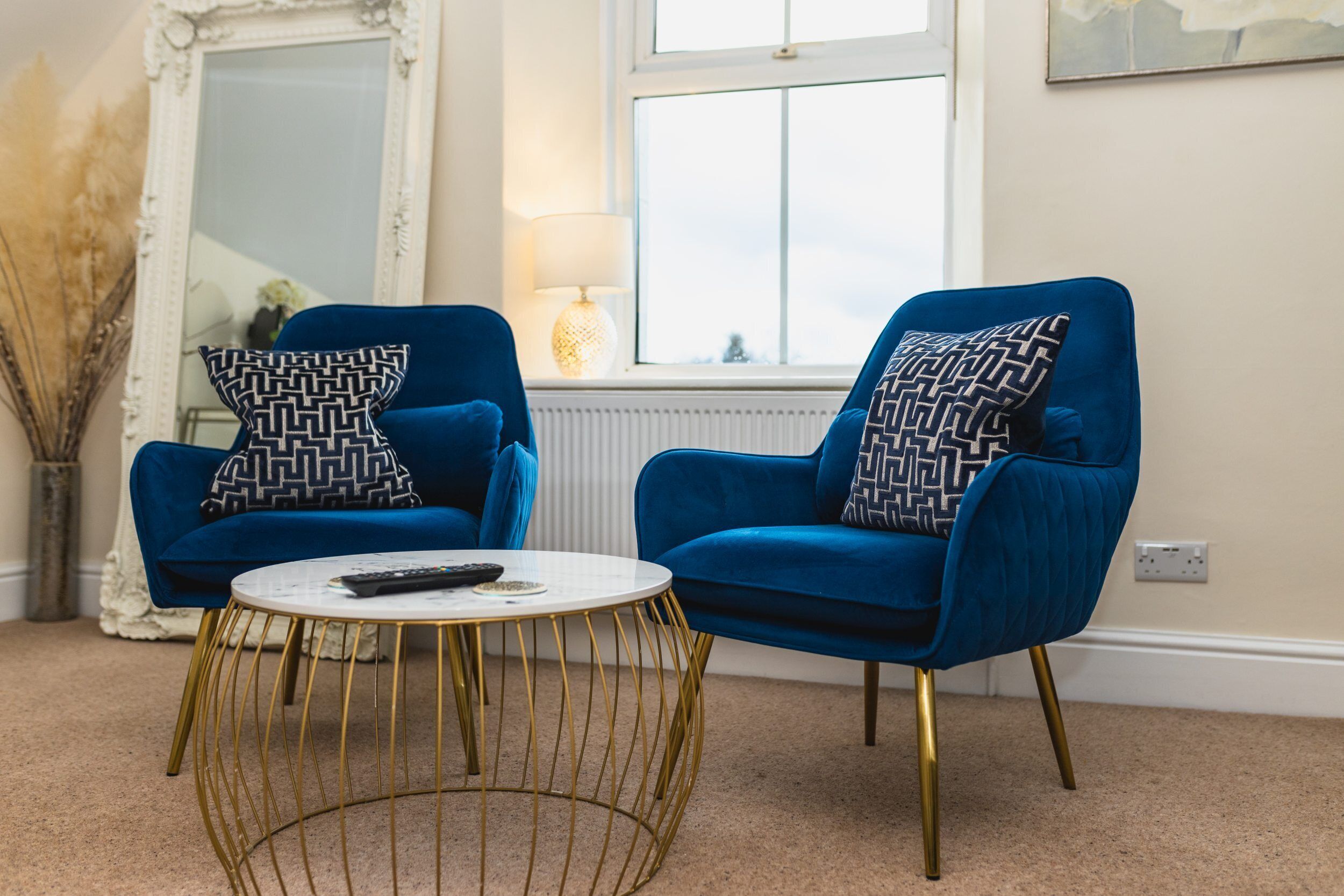 A living room with two blue armchairs, a round gold wire coffee table, a white lamp, and a large mirror.