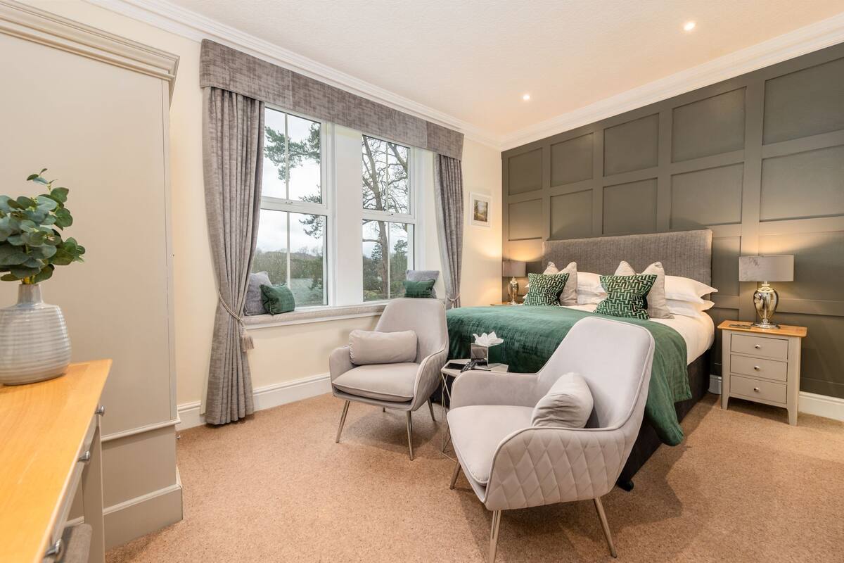 A bedroom with a large bed, two armchairs, a nightstand, a window with curtains, and a plant.