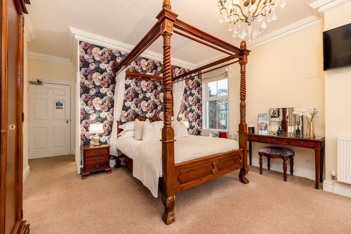 A four-poster bed in a room with floral wallpaper, a wooden desk, and a chandelier.