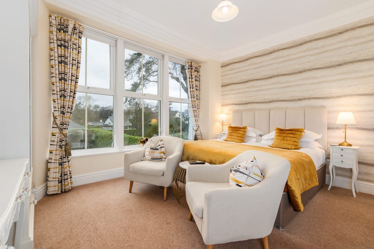 A bedroom with a large bed, two armchairs, a bedside table, and a view of the outdoors.