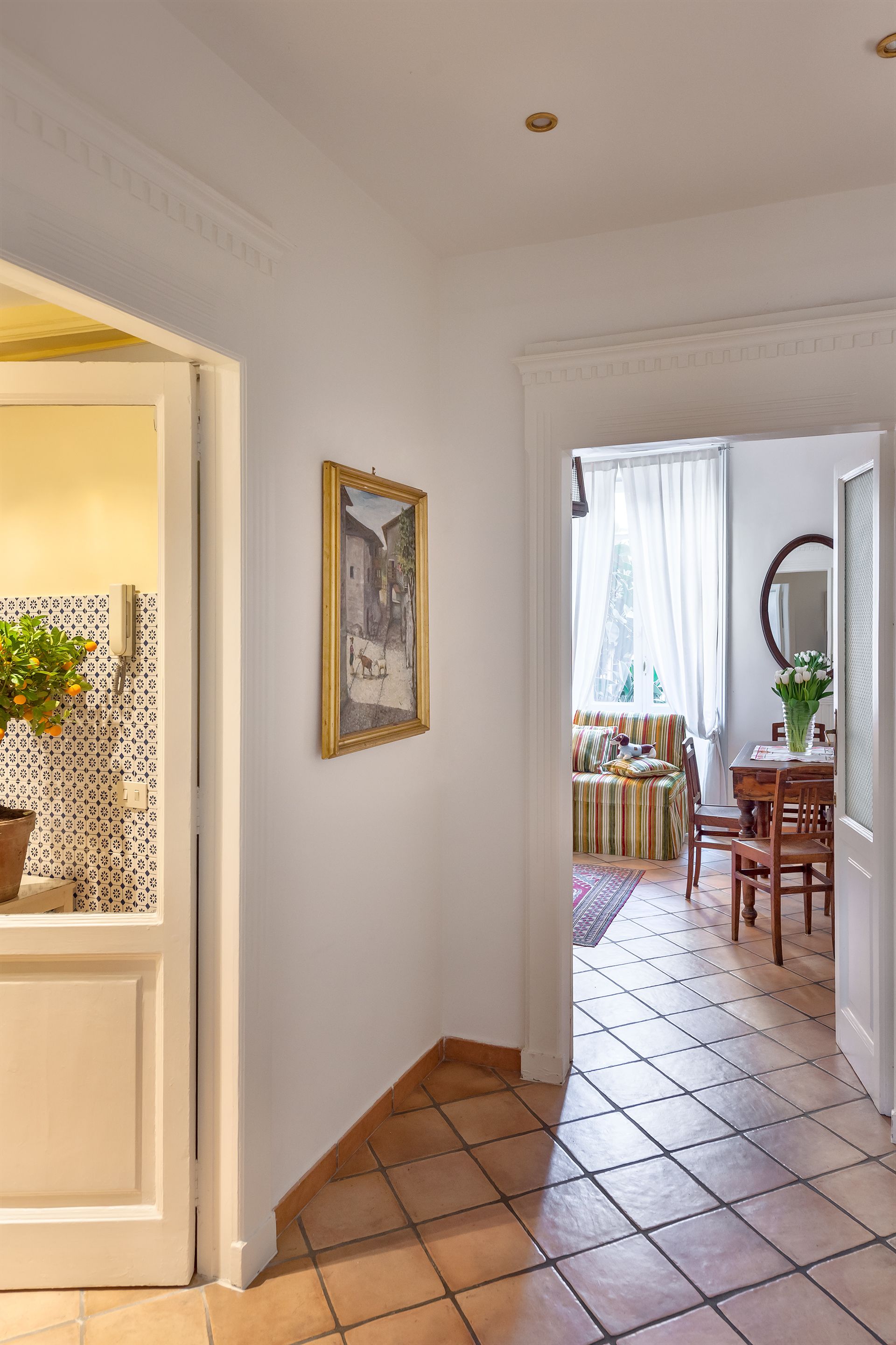 A hallway with a tiled floor, a door leading to a room with a patterned wall, a framed painting, and a dining area with a table and chairs.