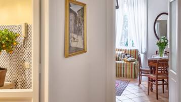 A hallway with a tiled floor, a door leading to a room with a patterned wall, a framed painting, and a dining area with a table and chairs.