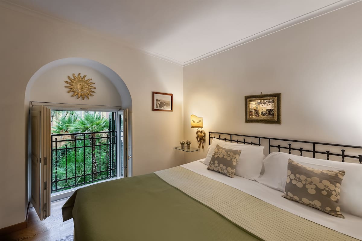 A bedroom with a bed, a nightstand, a lamp, and a window with a view of greenery.