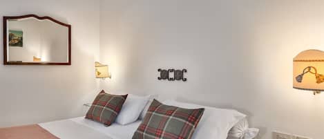A neatly made bed with plaid pillows, a mirror, wall-mounted lights, and a small shelf with plants.