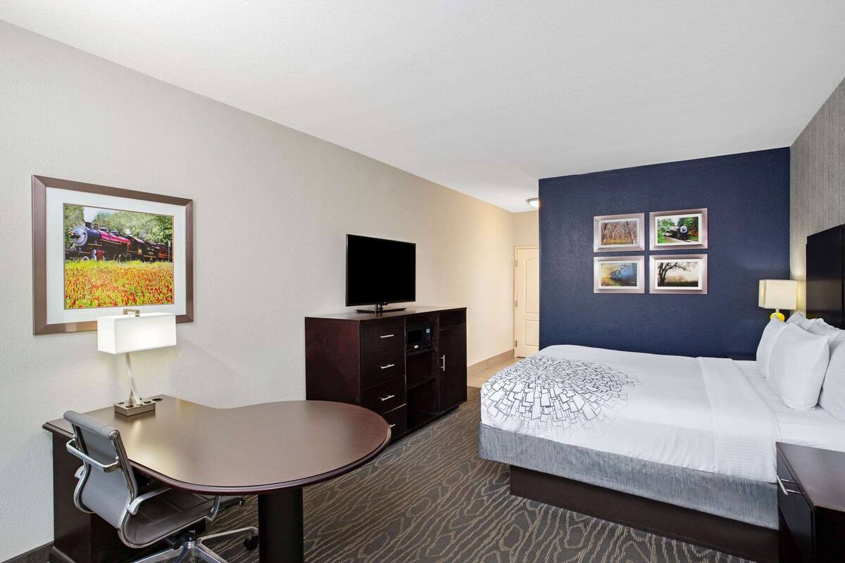 Ein Hotelzimmer mit einem Bett, einem Schreibtisch mit Stuhl, einem Fernseher und einer Wand mit vier gerahmten Bildern.
