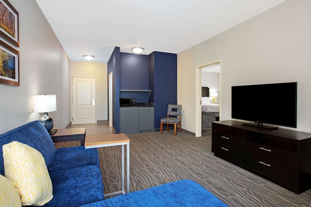 Ein Hotelzimmer mit einem blauen Sofa, einem Fernseher, einem kleinen Tisch, einem Sessel und einer Küchenzeile im Hintergrund.