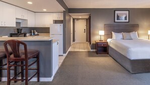 A modern hotel room with a bed, bedside tables, a desk, and a kitchenette.