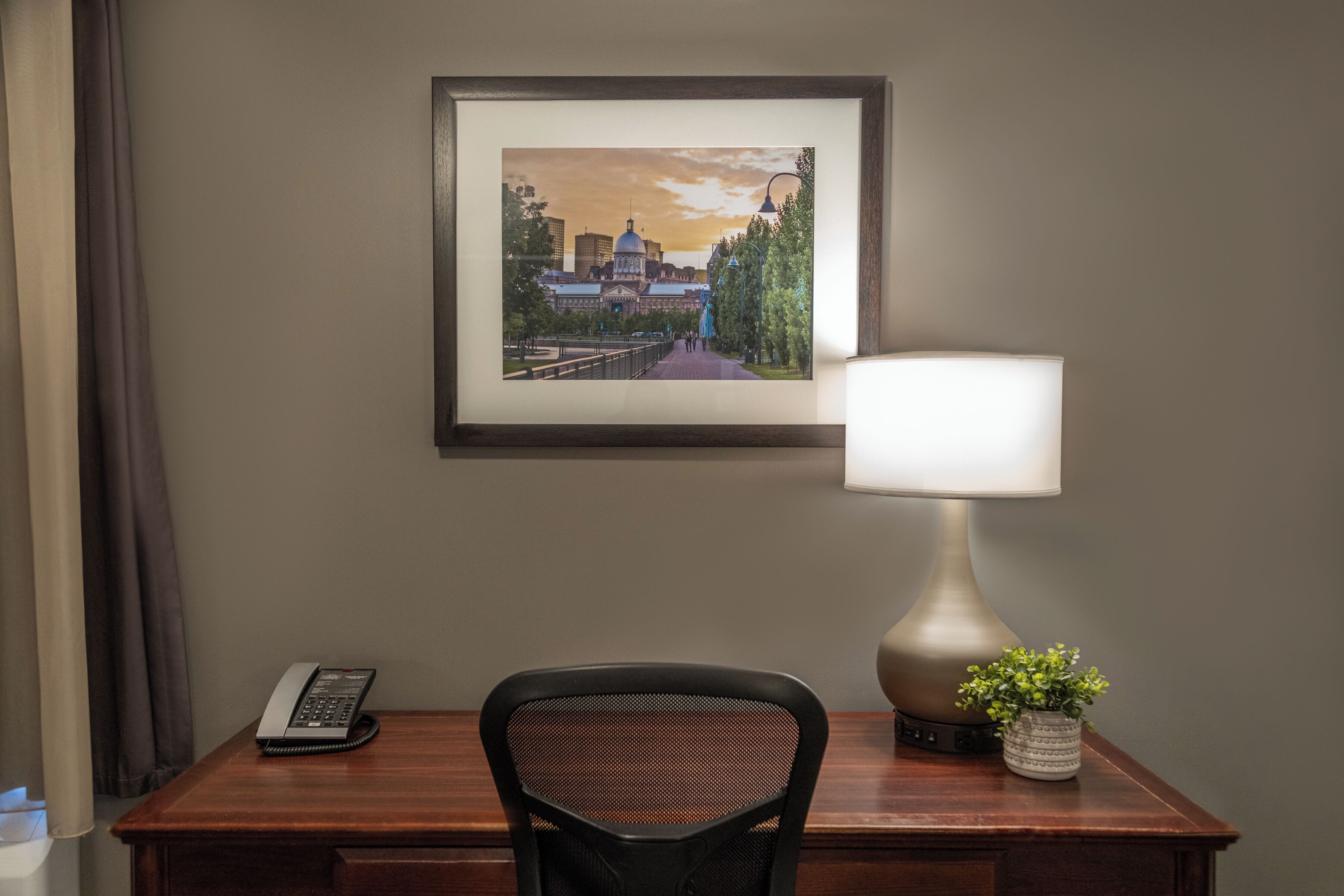 A hotel room with a desk, a framed cityscape picture, a lamp, and a telephone.
